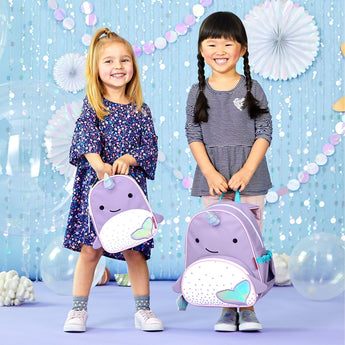 Two smiling toddlers hold purple Skip Hop Zoo Narwhal mini backpacks with iridescent fins.