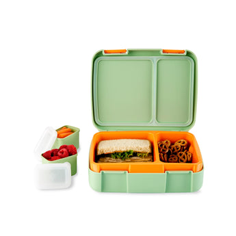 Open Skip Hop Zoo bento box with green exterior, orange divided tray holding a sandwich and pretzels. Two small green containers with raspberries and carrots sit beside it.