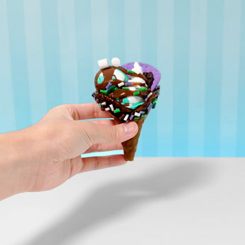 Hand holding a brown ice cream cone with chocolate, purple, and mint slime, sprinkles, and marshmallows.