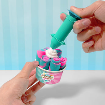 Hands adding white whipped cream slime to a cup of pink and blue rolled slime, against a striped blue background.