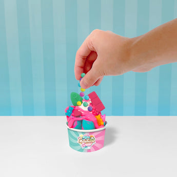 Hand adding colorful beads to a pink slime "gelato" in a branded cup with blue and pink accents.