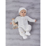 Newborn in white organic cotton Sophie la girafe outfit with matching hat, booties, and giraffe teether on a grey blanket.