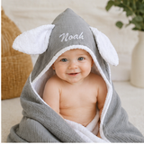Baby wrapped in a gray hooded towel with elephant ears, sitting on a couch.