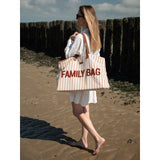 Woman walking on a beach, carrying a large striped beige and white canvas "FAMILY BAG" tote with brown leather straps.