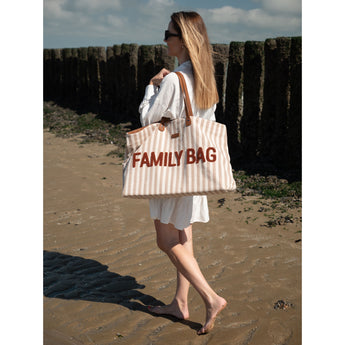 Woman walking on a beach, carrying a large striped beige and white canvas "FAMILY BAG" tote with brown leather straps.