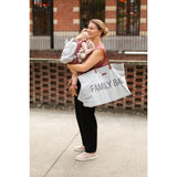Smiling mom holding a baby and a large light grey "FAMILY BAG" tote with brown leather handles.
