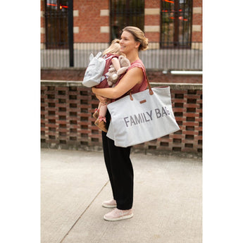Smiling mom holding a baby and a large light grey "FAMILY BAG" tote with brown leather handles.
