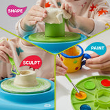 Child shaping clay on a green and blue motorized pottery wheel, sculpting with a tool, and painting a yellow clay mug.