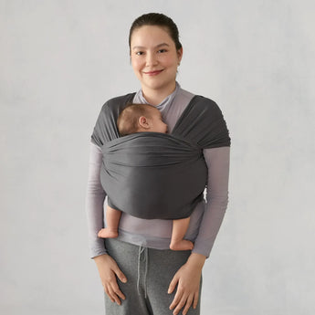 Smiling woman wearing a grey Ergobaby Aura knit wrap with a sleeping baby nestled inside.