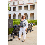 Woman in white jumpsuit carrying baby in a gray Ergobaby Away carrier, standing near strollers.