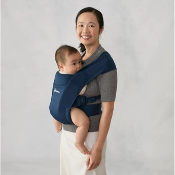 Smiling woman wearing a navy Ergobaby Embrace newborn carrier with a baby facing inward.