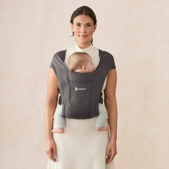 Woman smiling, wearing a heather grey Ergobaby Embrace newborn carrier with a baby sleeping in front.