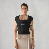 Woman smiling, wearing a black Ergobaby Embrace newborn carrier with a sleeping baby.