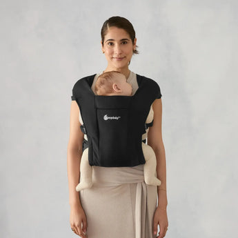 Woman smiling, wearing a black Ergobaby Embrace newborn carrier with a sleeping baby.