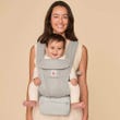 Smiling woman carrying a happy baby in a light gray Ergobaby Omni 360 carrier.