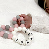 Baby's hand holding a personalized silicone teether with pink, grey, and white beads spelling "JUMANA".