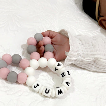 Baby's hand holding a personalized silicone teether with pink, grey, and white beads spelling "JUMANA".