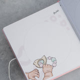 Open pink suede baby milestone album page with "التاريخ" (Date) and baby hands holding a pastel rattle illustration.