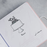 Open pink baby milestones photo album page with "is One" cake illustration and Arabic text.