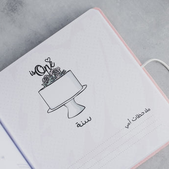 Open pink baby milestones photo album page with "is One" cake illustration and Arabic text.