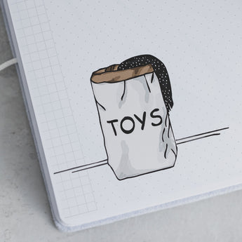 Hand-drawn illustration of a white paper bag labeled "TOYS" with a black polka-dot fabric peeking out, on a grid notebook page.