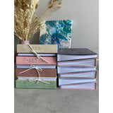 Stack of personalized suede baby milestone photo albums in beige, pink, brown, and green, with a dark gray album open.