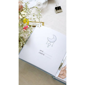Open baby blue personalized milestone photo album with a moon and stars design, showing name/surname lines.