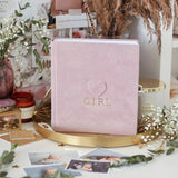 Pink suede baby milestone photo album with "GIRL" and heart embossed in gold, on a gold tray.