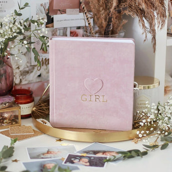 Pink suede baby milestone photo album with "GIRL" and heart embossed in gold, on a gold tray.