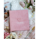 Pink suede baby milestones photo album with "GIRL" and heart embossed, surrounded by flowers and baby photos.