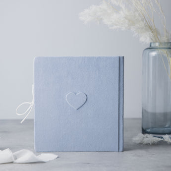Light blue suede baby milestones photo album with an embossed heart, white ribbon, and a blue glass vase.