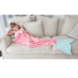 Child in a pink mermaid sleeping sack with a light blue tail, relaxing on a cream sofa.