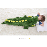 Green crocodile-shaped kids sleeping sack with yellow accents, a child sleeping inside on a white fluffy rug.