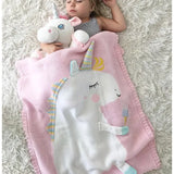 Sleeping child with a pink knit unicorn blanket and a white unicorn plush toy on a fluffy white rug.