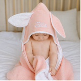 Personalized organic peach bunny hooded towel with white trim, embroidered "Ella" on the hood, worn by a baby.