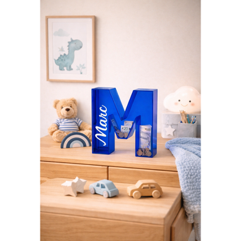 Blue acrylic letter "M" piggy bank with "Marc" printed in white, holding coins and bills.