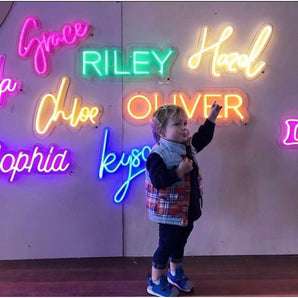 Child pointing at colorful custom LED neon name signs: Grace (pink), Riley (green), Hazel (yellow), Chloe (orange), Oliver (orange), Sophia (pink), Kyson (blue).