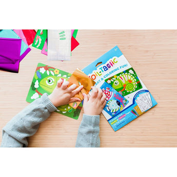 Child's hands applying gold foil to a green chameleon design from the Ooly Foil-Tastic art kit on a wooden table.