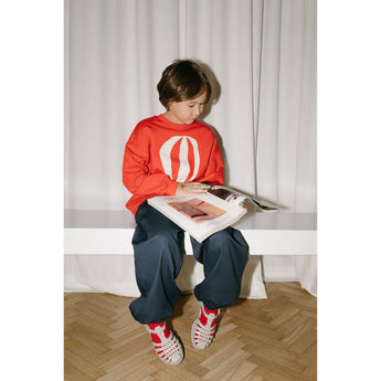 Kid in an oversized red organic cotton sweatshirt with a white hot air balloon print, blue pants, and red and white sandals.