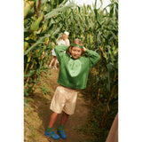 Kid in an oversized green organic cotton sweatshirt with a light green potato face print, in a cornfield.