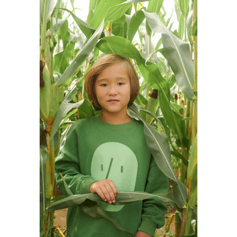 Kid in an oversized green organic cotton sweatshirt with a light green potato face print, standing in a cornfield.