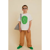 Kid in beige organic cotton Potato Face t-shirt with green print, brown pants, and green sandals.