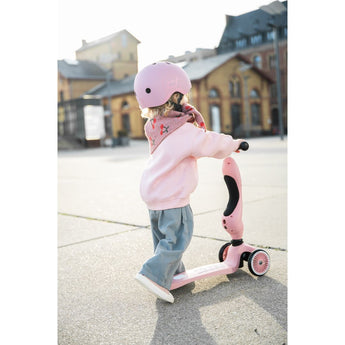 Toddler in pink helmet and outfit pushing a pink Scoot & Ride Highwaykick 1 scooter outdoors.