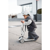Toddler in a white helmet and gray vest riding a white Scoot & Ride Highwaykick 1 scooter outdoors.
