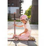 Young girl in pink helmet and checkered overalls adjusting a pink Scoot & Ride Highwaykick 3S LED scooter.