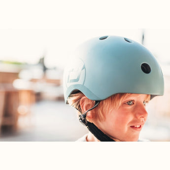 Blonde child wearing a light blue Scoot & Ride eco-friendly kids helmet with a white logo and black strap.
