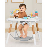 Baby in Skip Hop Discoverosity 3 Stage Activity Center with white table, wooden legs, and colorful toys.
