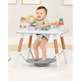 Baby in Skip Hop Discoverosity 3 Stage Activity Center with white table, wooden legs, and colorful toys.
