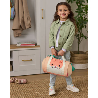 Smiling child holds a peach Skip Hop Duffimals cat duffle bag with expandable sides and green handles.