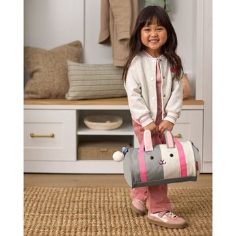 Smiling child holds a gray, white, and pink Skip Hop Duffimals bunny duffle bag with expandable sides.